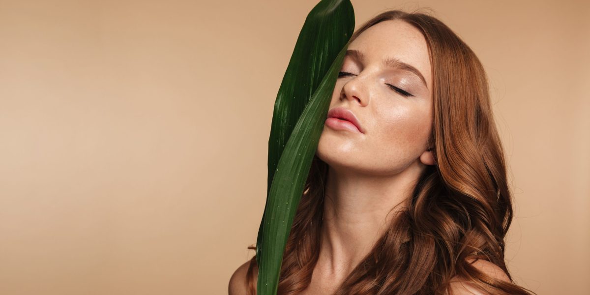 Beauty portrait of ginger woman with long hair posing with green leaf over cream background